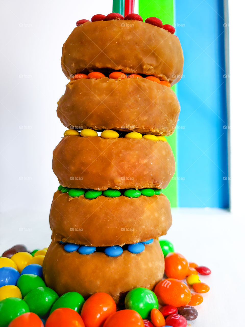 Five stacked donuts decorated with M&M minis and surrounded by peanut M&Ms on a white surface with a blue, green and white solid background.  Reminiscent of the classic Fisher Price rock-a-stack infant toy.