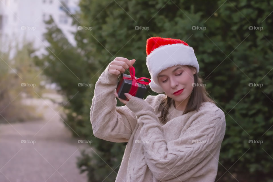 Close up portrait of happy young cute woman  in a cozy knitted beige sweater in a santa claus hat holding gift in red packaging with bow near her ear, she wants to know what inside of it, standing on the street outside near the Christmas trees