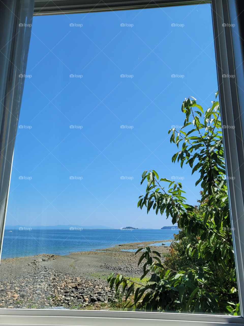 Looking out clear glass window to sunny beach view with blue sky and ocean, with sand and rocks