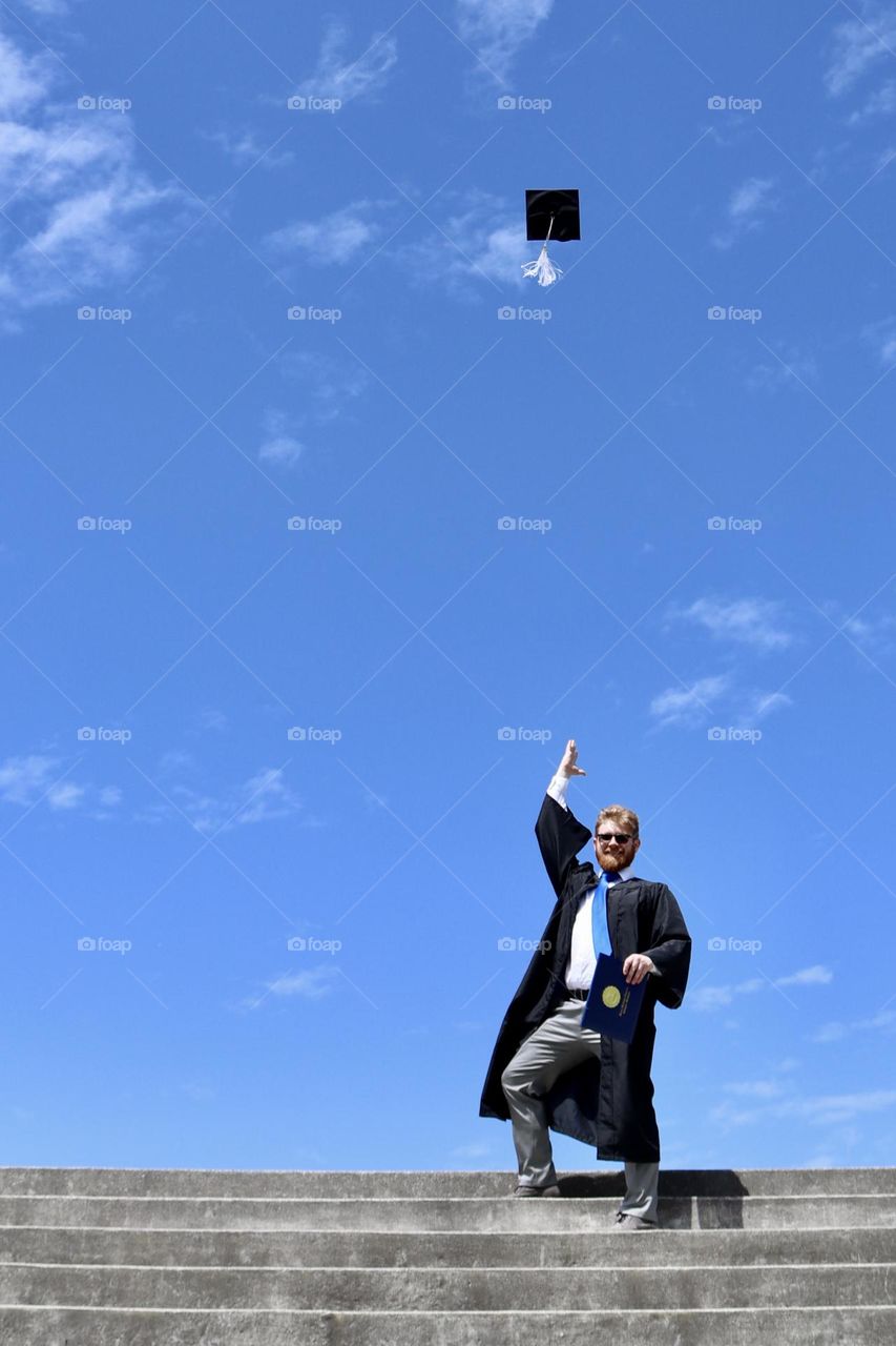 A recent college graduate throws his black graduation cap in the air to celebrate, blue sky framing the shot