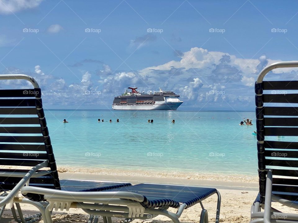 Lounge chairs on the beach with people swimming in front of a cruise ship