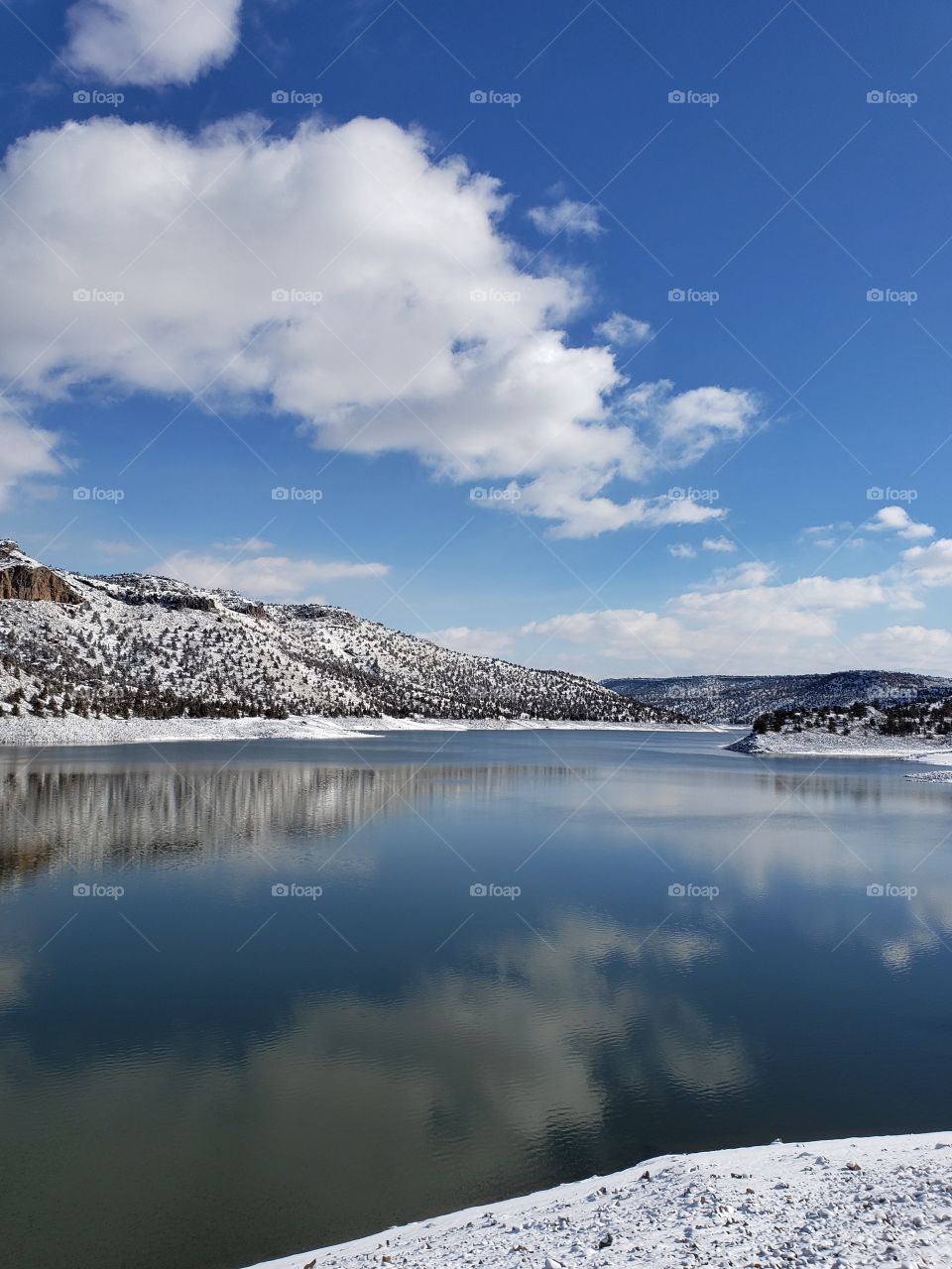 Snow covered hills, trees, clouds, and a bright blue sky reflect in the partially iced over Prineville Reservoir on a sunny winter day in Central Oregon. 