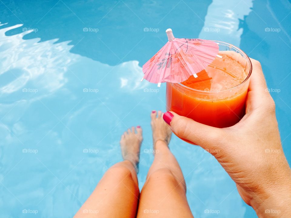Woman hand with pink nail polish holding glass of orange juice with umbrella over the pool blue water