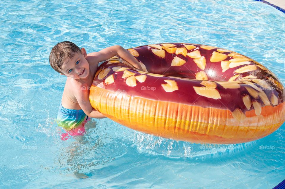 Little boy having fun playing with inflatable big donut toy in swimming pool.