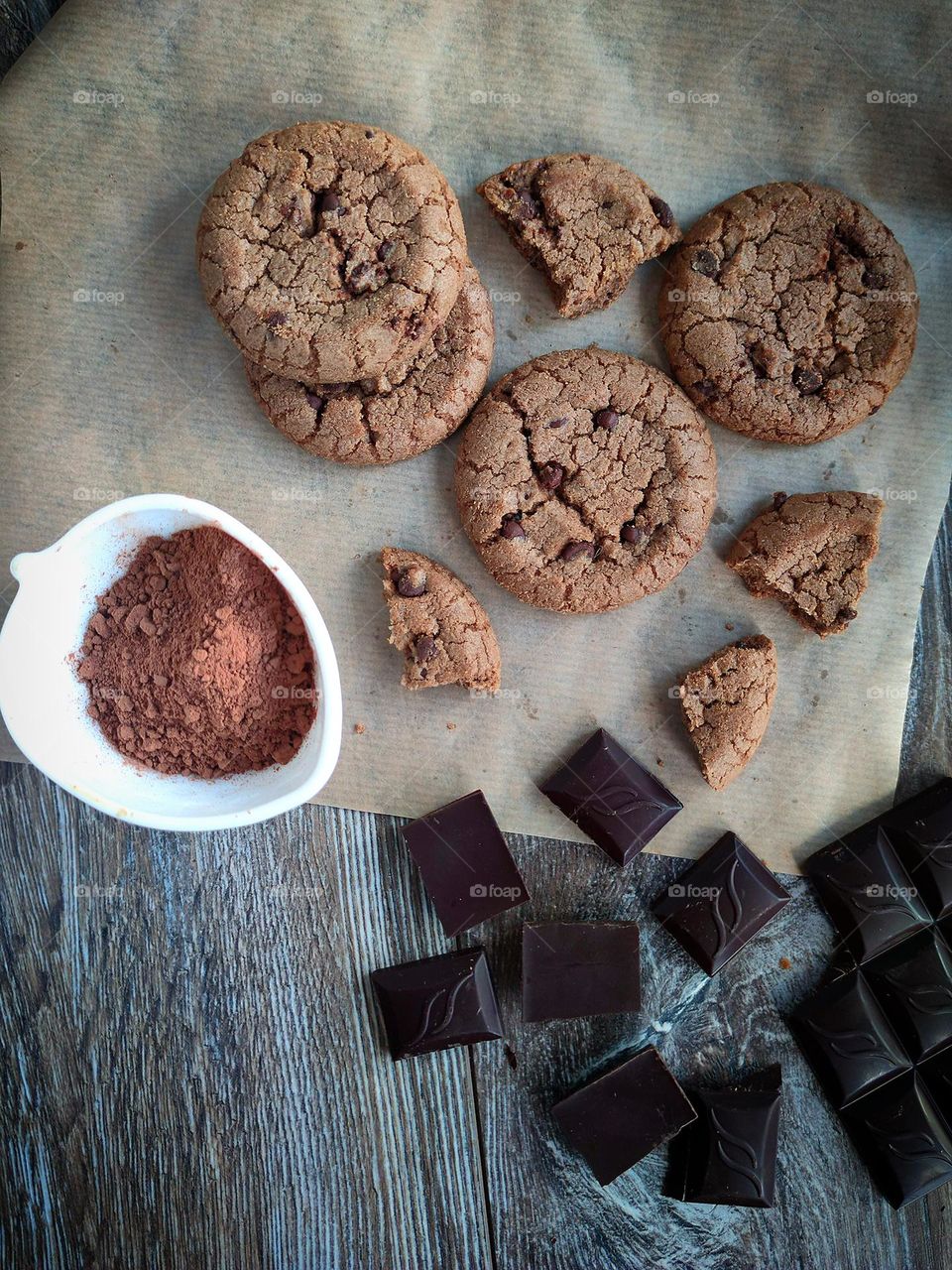 On a wooden background lie: baking paper on which chocolate cookies lie, a white cup with cocoa powder and pieces of dark chocolate.View from above