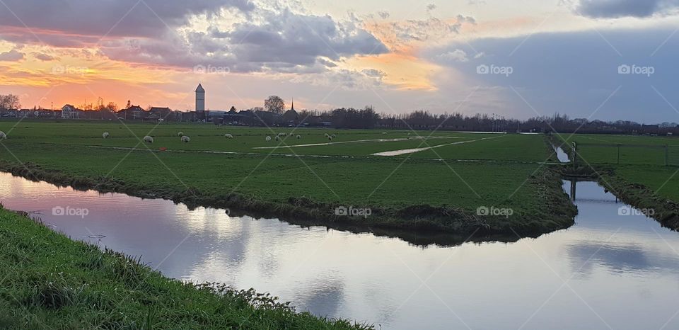 Dutch landscape at sunset with view over the fields and ditches , colorful clouds in the sky and a watertower at the horizon