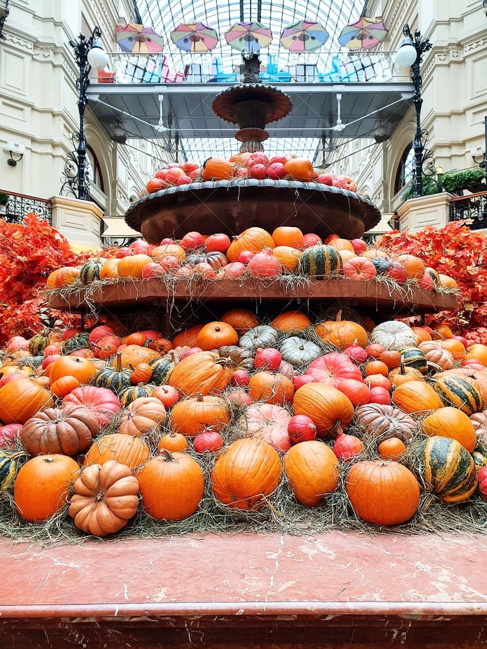 A fountain made of different varieties of pumpkin.  Autumn colors.  There are umbrellas in the background
