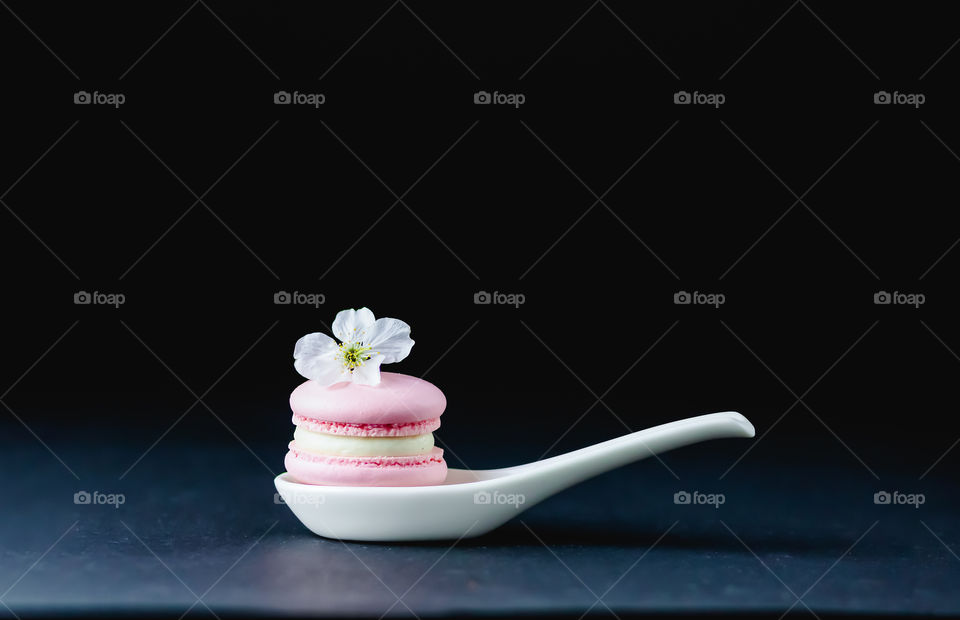 Still life made by pink macarons with cream, cherry flower and white spoon on the dark background.