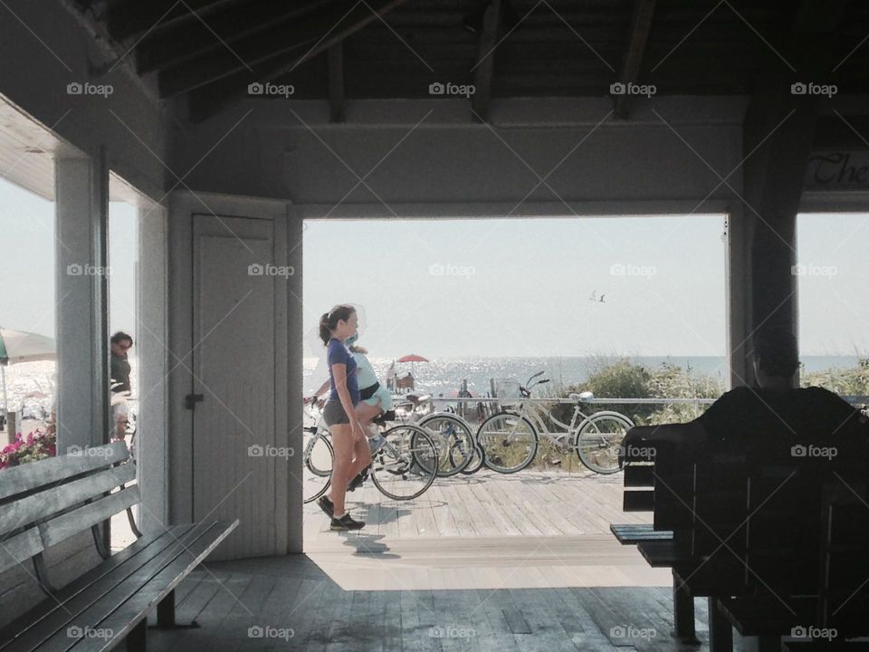 People walk and ride bicycles on the Ocean Grove boardwalk, photographed from the open-sided Beach Pavilion, where church services are frequently held. The Pavilion offers shade and a beautiful view of the ocean, beach and passerby. 