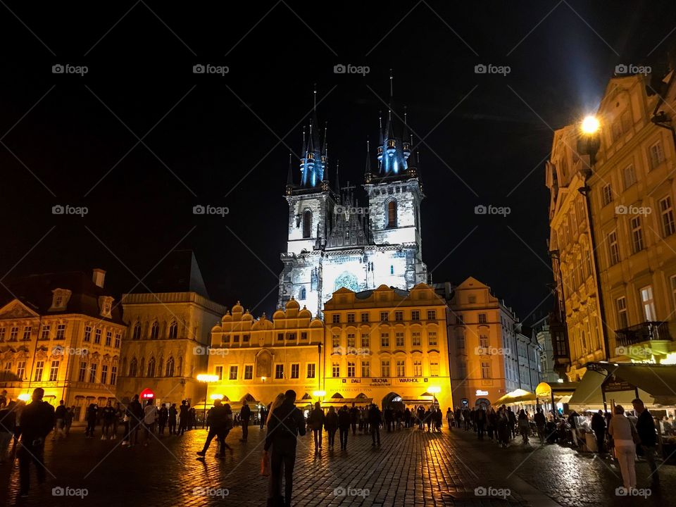 the square in the evening have a lot of people, buildings with the yellow light of reflective lights. Behind is a church illuminated by blue lights, look really outstanding against the black sky background