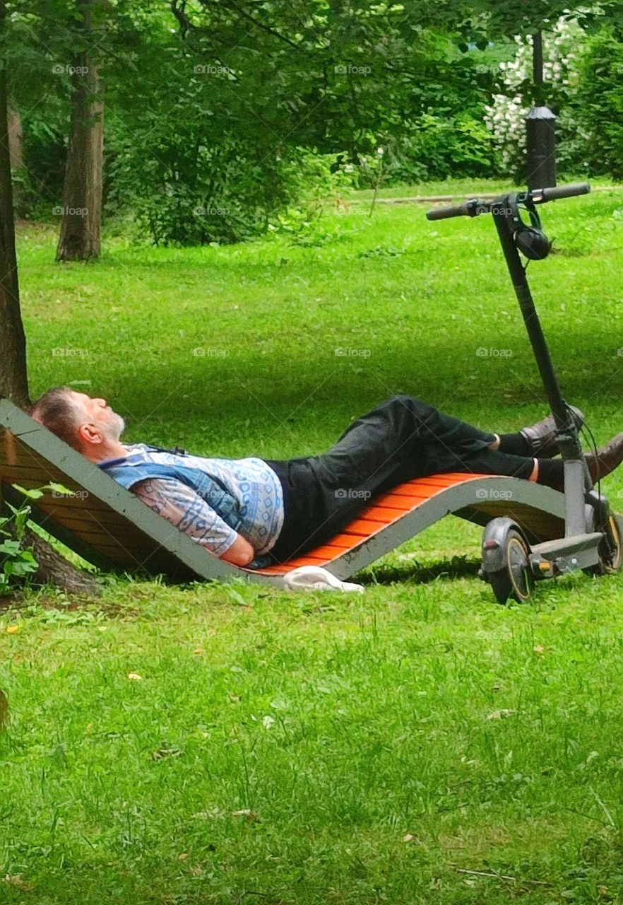 A park.  On the green grass among the trees, a man lies on a wooden deck chair and relaxes.  Nearby is an electric scooter