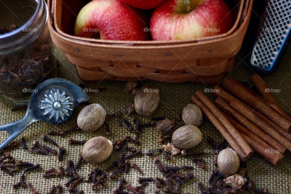 Cinnamon sticks, whole nutmeg, whole cloves, a vintage pie crust crimper, a small glass jar, apples in a basket, and a spice grater on burlap