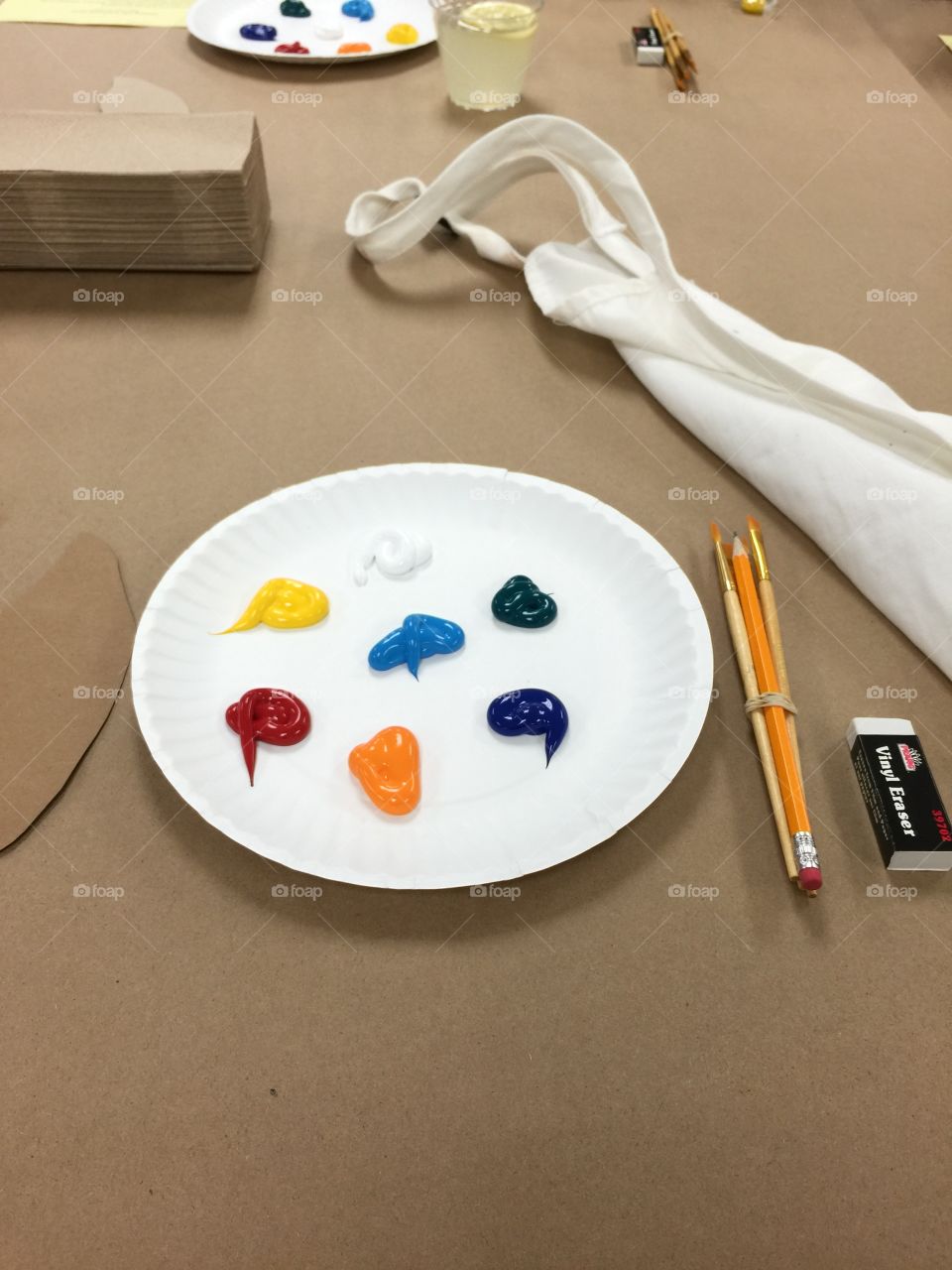 Setup for oil painting with palette, brushes, eraser, pencil, and apron on top of a craft paper covered table