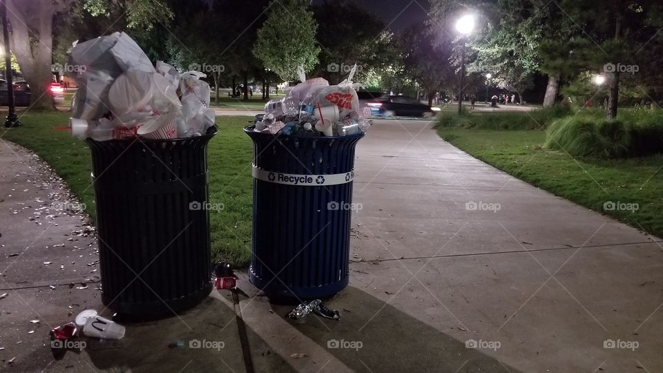 A trash can and recycling bin overflowing with trash in a public park.