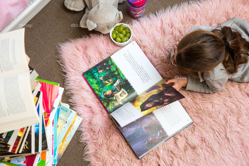 Little girl reading in her bedroom with healthy snacks on the side. A big stack of books show her love for reading and knowledge. Staying in good shape is about creating good habits from a young age.