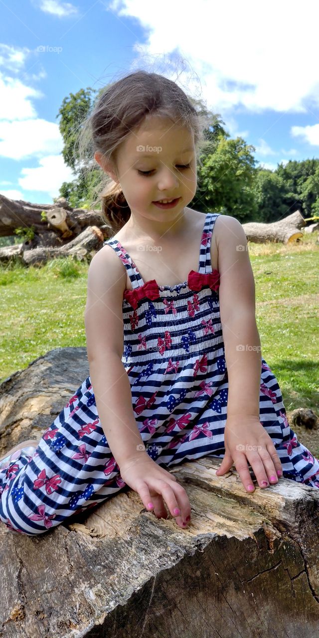 child in dress in summer sat on log smiling looking thoughtful