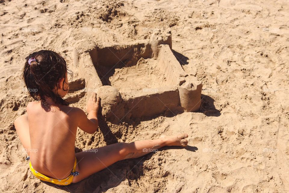 One little caucasian girl sitting with her back builds a sand castle on the shores of the Boliarian Sea on a clear sunny day in spain, close-up side view.