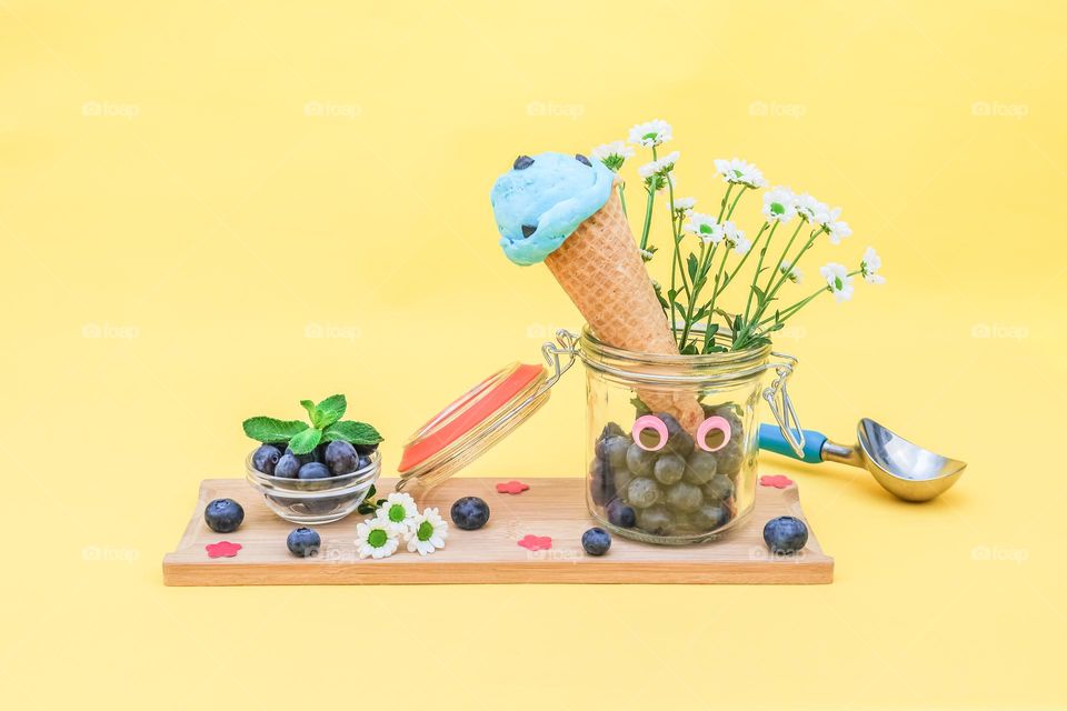 Blueberry ice cream in a waffle cone stands in a glass jar with fresh berries and flowers on a wooden cutting board on a yellow ion, close-up side view. Concept summer treat, summer food, natural ice cream, summer time, summer mood, healthy lifestyle