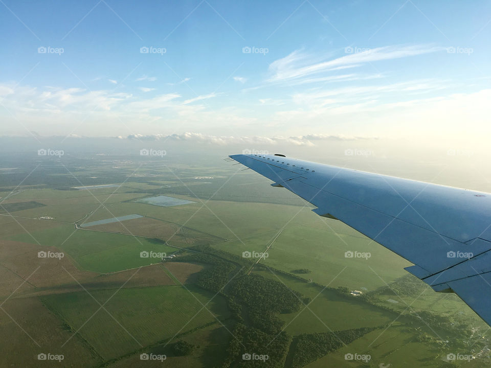 Airplane wing flying over green fields sunny evening