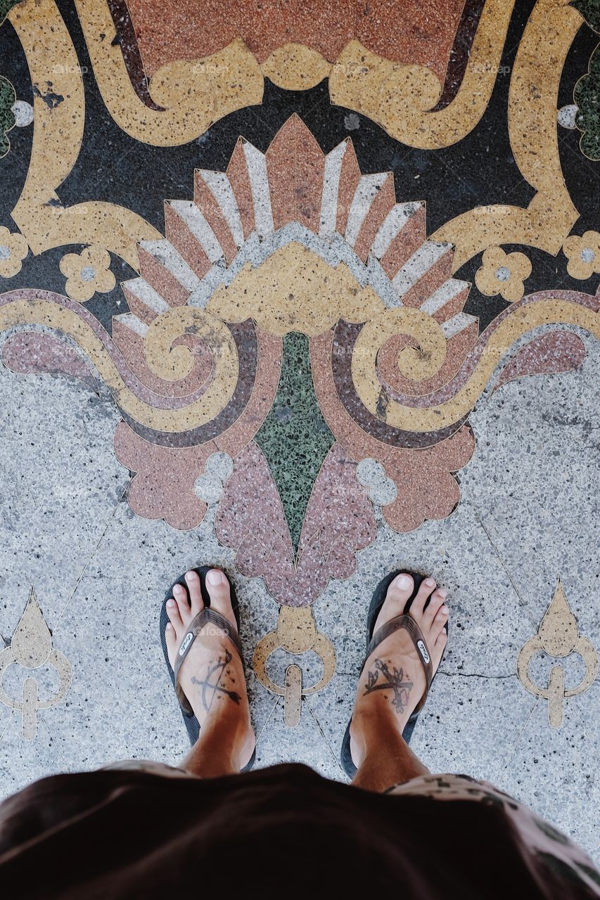 Tattooed feet in sandals standing in front of some intricate floor tile design. 