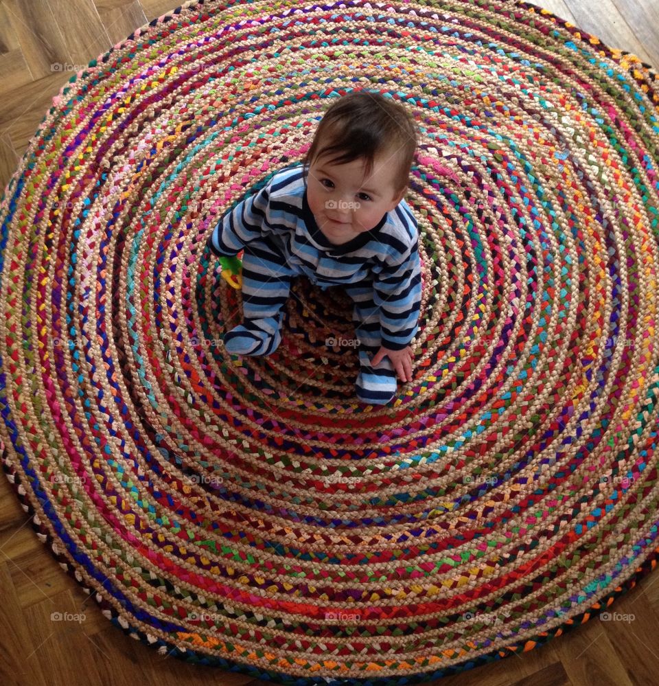 Baby sitting in the middle of a colourful circle. Sitting on a rug looking up at the camera.