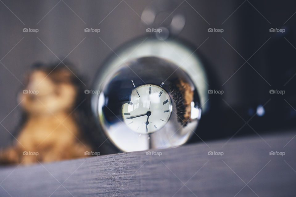 A portrait of a clock shot through a glass lens ball. the clock is behind the ball and its mirror image is being warped and is upside down inside of the ball.