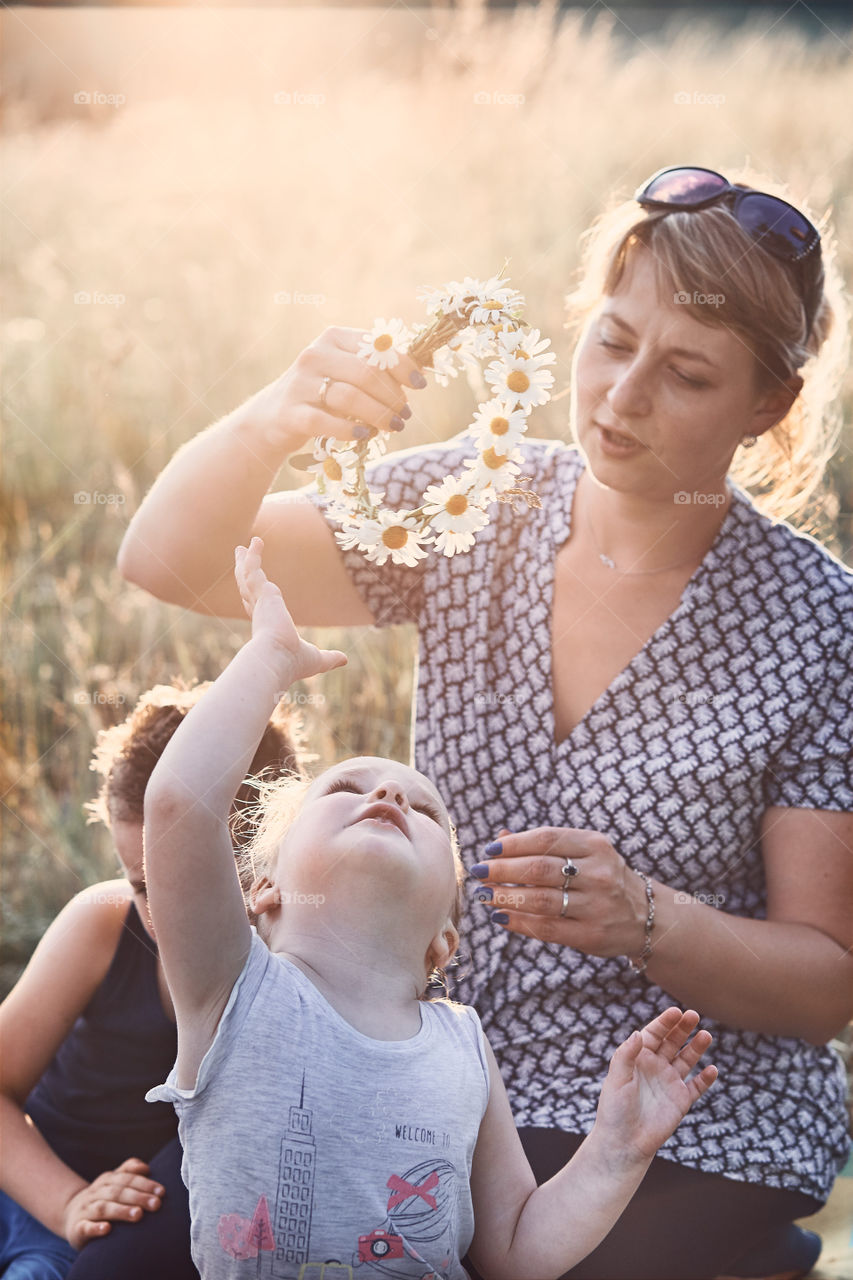 Mother putting a coronet of wild flowers on a head of little girl. Family spending time together on a meadow, close to nature. Parents and children playing together. Candid people, real moments, authentic situations