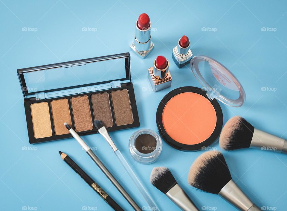 Makeup set of pallet of nude eyeshadow, round face powder, red lipstick, soft black eyebrow shadow, makeup brushes and eyebrow pencil on blue background, flat layered close-up. Concept female cosmetics, beauty salon.