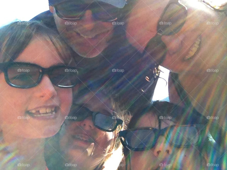 Selfie of my husband & I with our 3 daughters at the beach wearing our bathing suits & our 3D movie glasses.  The sun is peeking over my shoulder & made a beautiful array of colourful sunbeams in front of all our faces! 🌈
