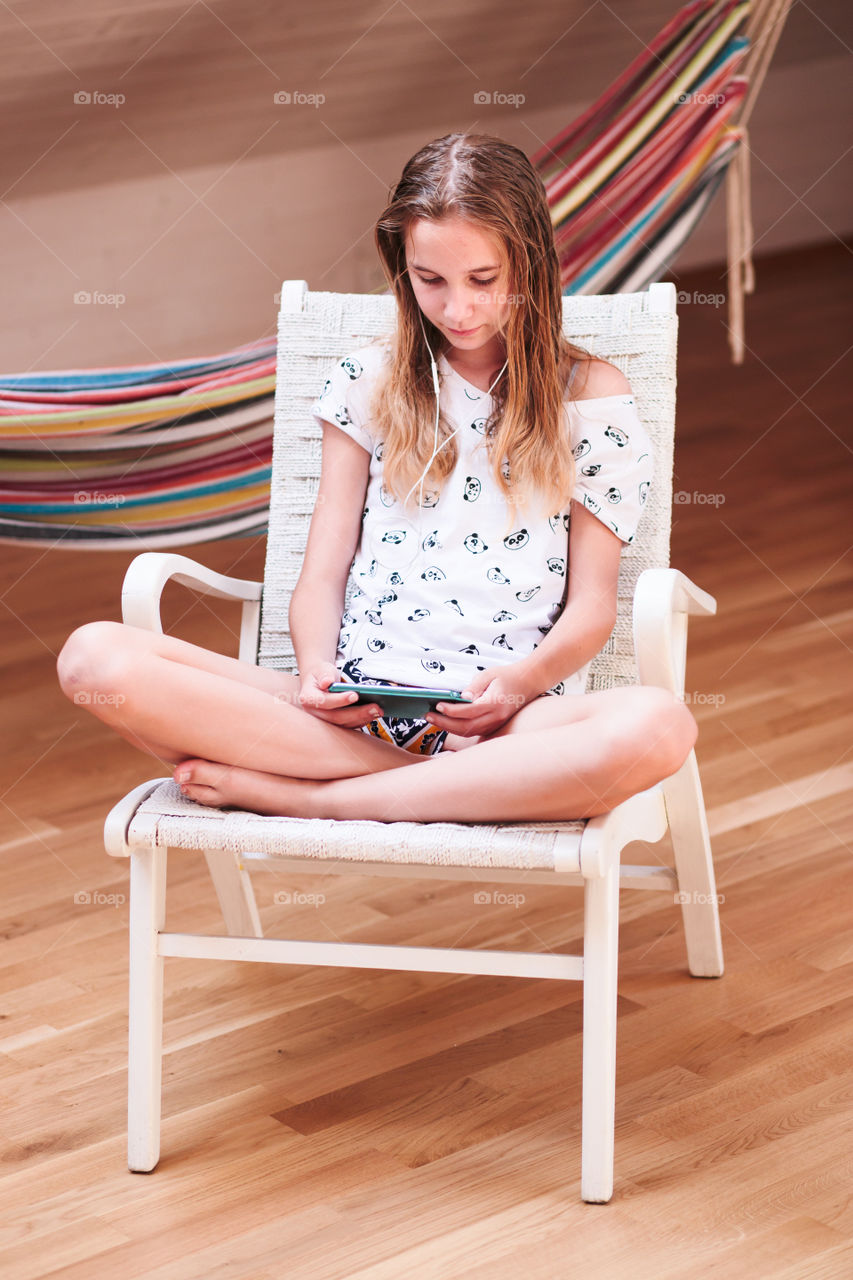 Teenage girl listening to music and shopping online on mobile phone sitting on chair at home