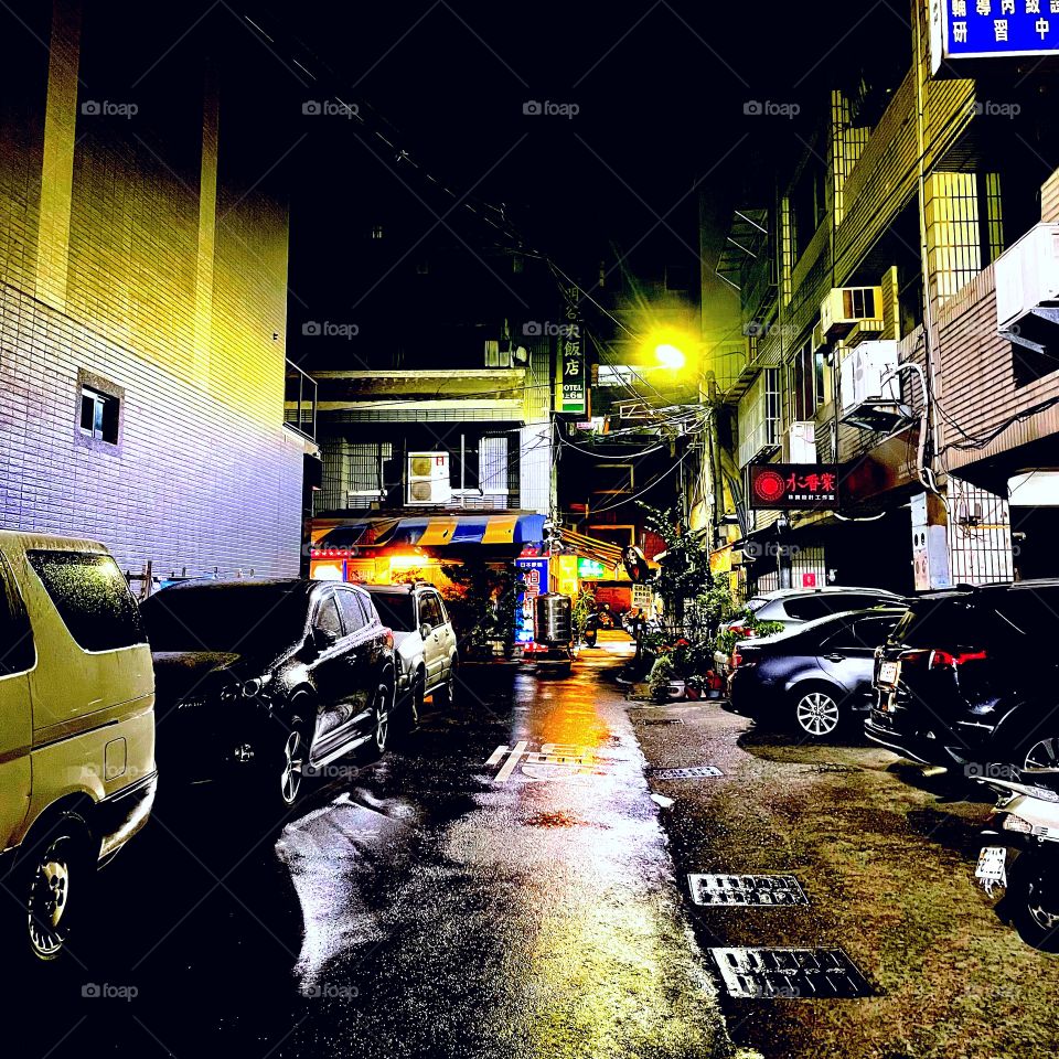 A dark alley on a rainy night. Several cars, scooters, businesses, and apartments line either side. 