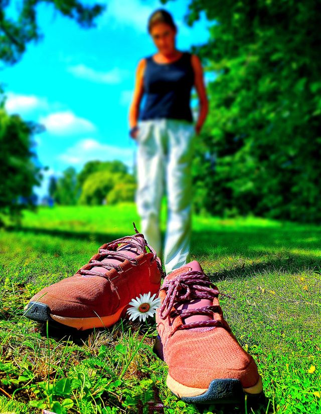Spring. Burgundy sneakers stand on the green grass near a daisy. In the background is a silhouette of a woman