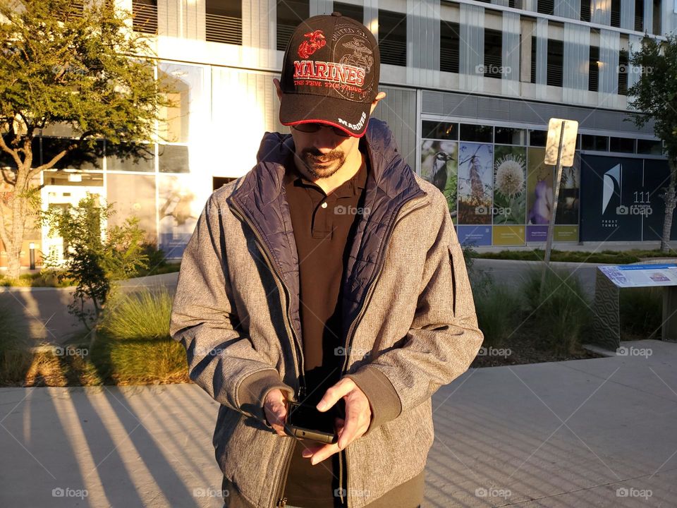 Demi saison. A man wearing a light gray jacket , a black polo shirt, denim blue jeans and a black cap. He is standing in front of a city building while using his phone at sunset. A casual fall season look while strolling downtown.