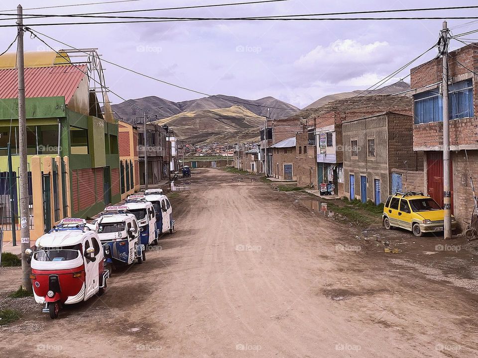 Red and blue mototaxis parked in line along dusty street and houses with mountains in the background.