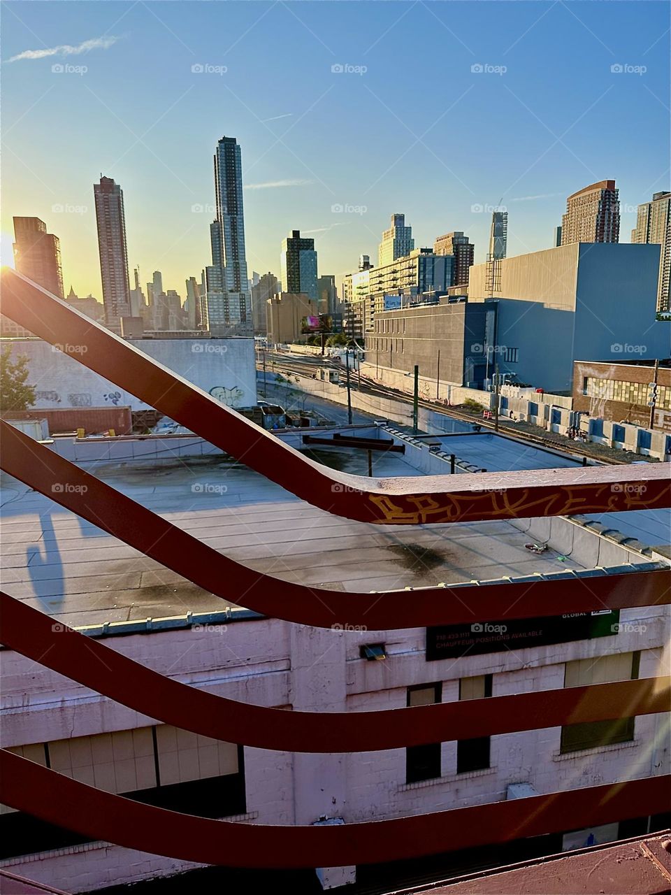 This is the red metal staircase of the “Pulaski Bridge” that connects “Greenpoint”, Bklyn to LIC overlooking the “Long Island Railroad” train tracks and “iFly”, a large rectangular building with an indoor flight simulator. 2024. Hypnotic Productions