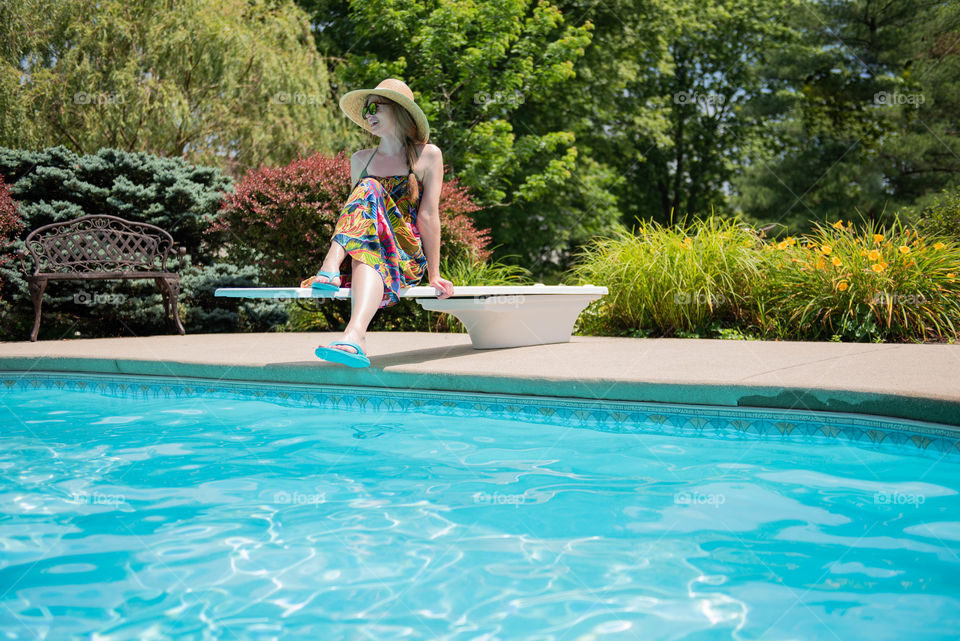 Young millennial woman wearing a dress and flip flops while sitting on the diving board of a swimming pool in the summer