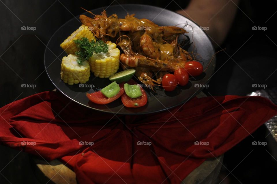 Asian food balado fried prawns with corn chunks, lemon slices and tomatoes served on black plate with red cloth bottom and wooden coaster. Asian style street seafood.