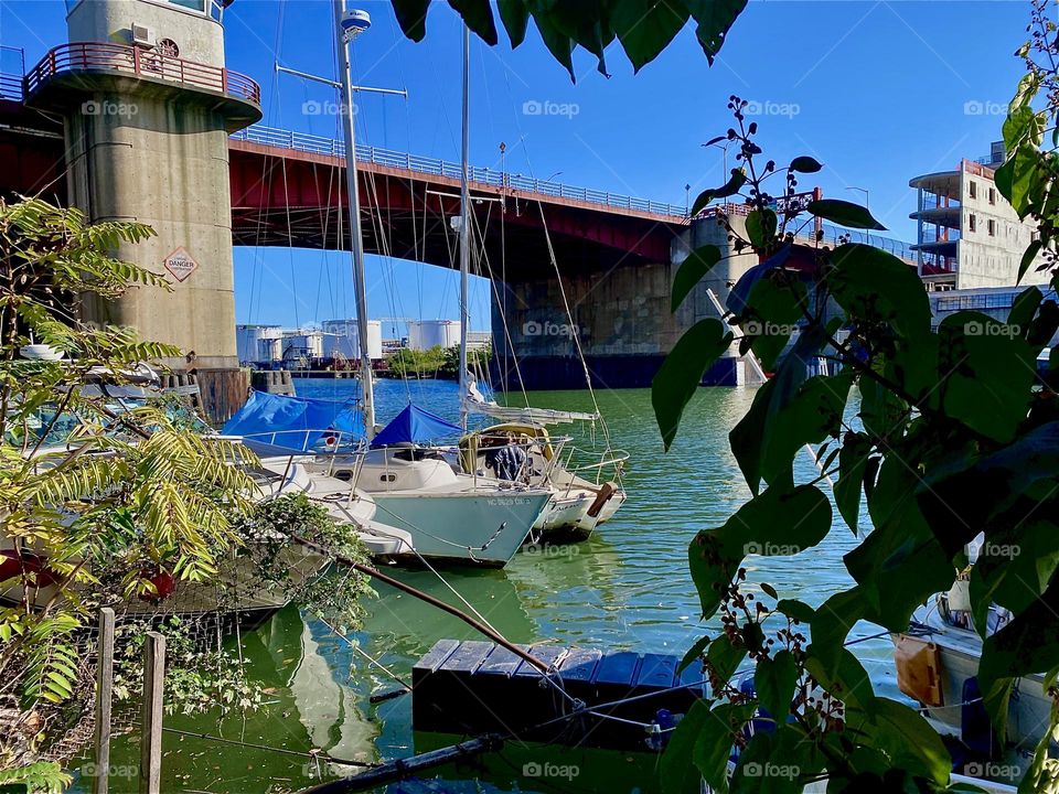 This is the “Pulaski Bridge” at “Newtown Creek” in LIC, Queens seen through the foliage of bushes on the narrow strip of land on the LIC shore. Two sailboats can also be seen. 2023. Hypnotic Productions