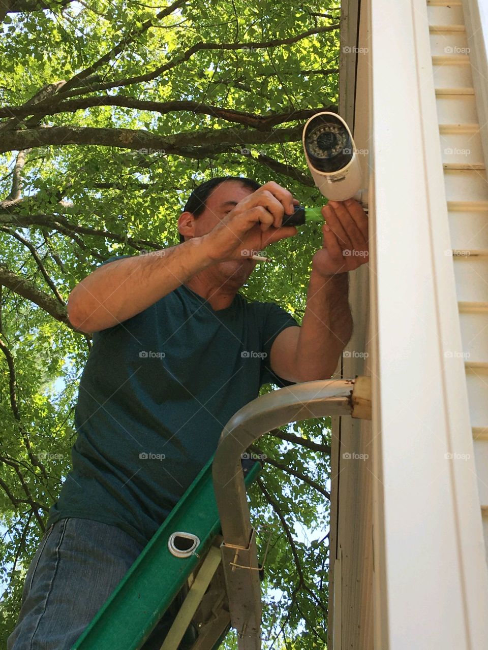 Man high up on ladder adjusting new camera to surveillance system in the home. Tools are being used getting angle right.
