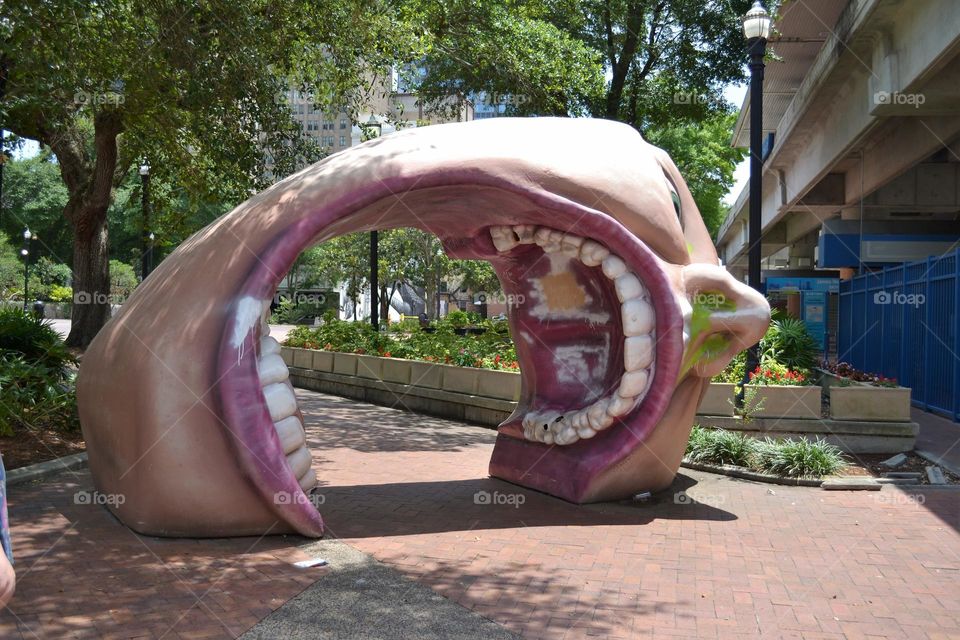 A sculpture of a huge head with the mouth gaping open showing teeth on a pathway in a park