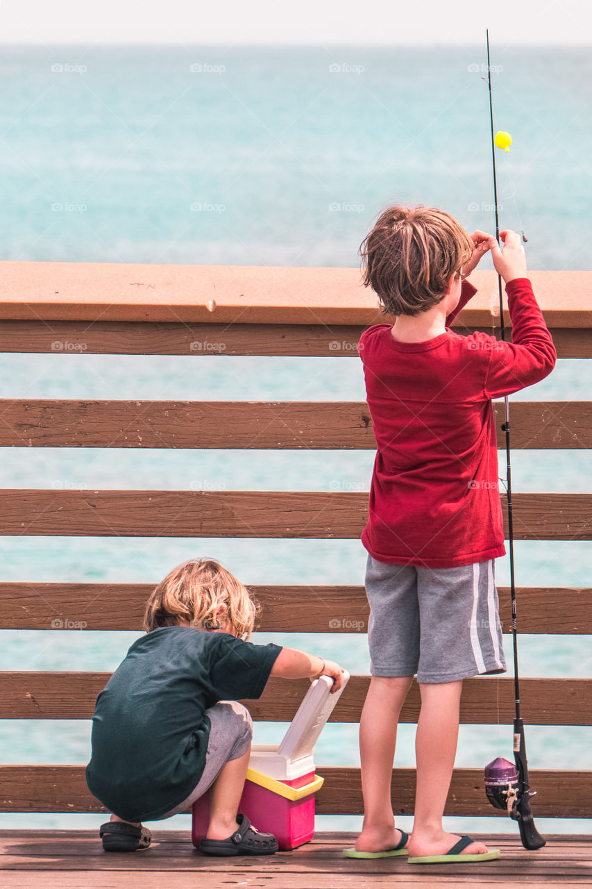 Two young boys maybe brothers help each other to prepare for fishing off a dock by the ocean on a beautiful sunny day with a bait box and fishing poll as their favorite hobby takes from a young age