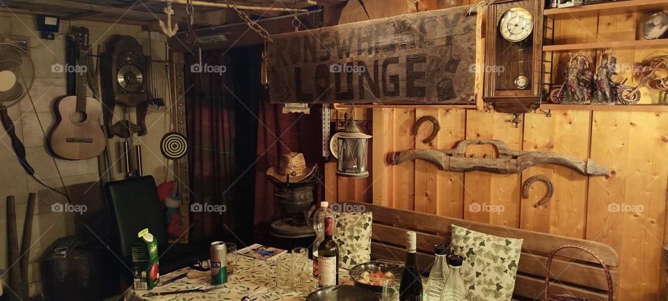 The other side of the interior of “Ron’s Whiskey Lounge” shows a wooden bench with pillows, a table with tablecloth, two bottles of wine, a can of beer and an ash tray. It’s a private club that obviously has been lived in. 2023. Hypnotic Productions