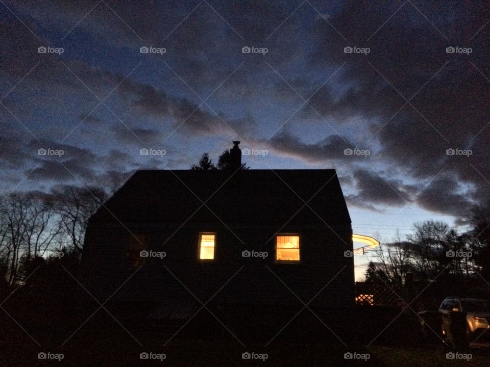 House at dusk. The sun sets behind a house with lights on.  
