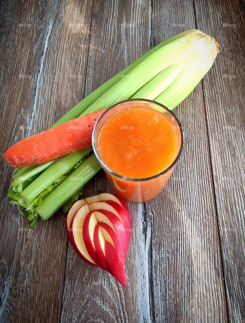 There is a glass with an orange smoothie on a wooden surface. Ingredients lie nearby: a stalk of celery, a carrot and a red apple, which is cut into a heart shape