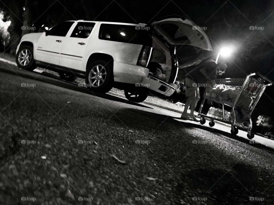 After The Grocery Shopping At Night, Transfering The Groceries From The Cart In The Back Of The Vehicle White SUV In The Parking Lot With Street Lights.