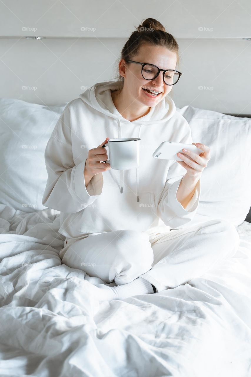 Hipster millennial Woman watching films on phone laughing holding phone relaxing in the bed holding white cup of coffee 