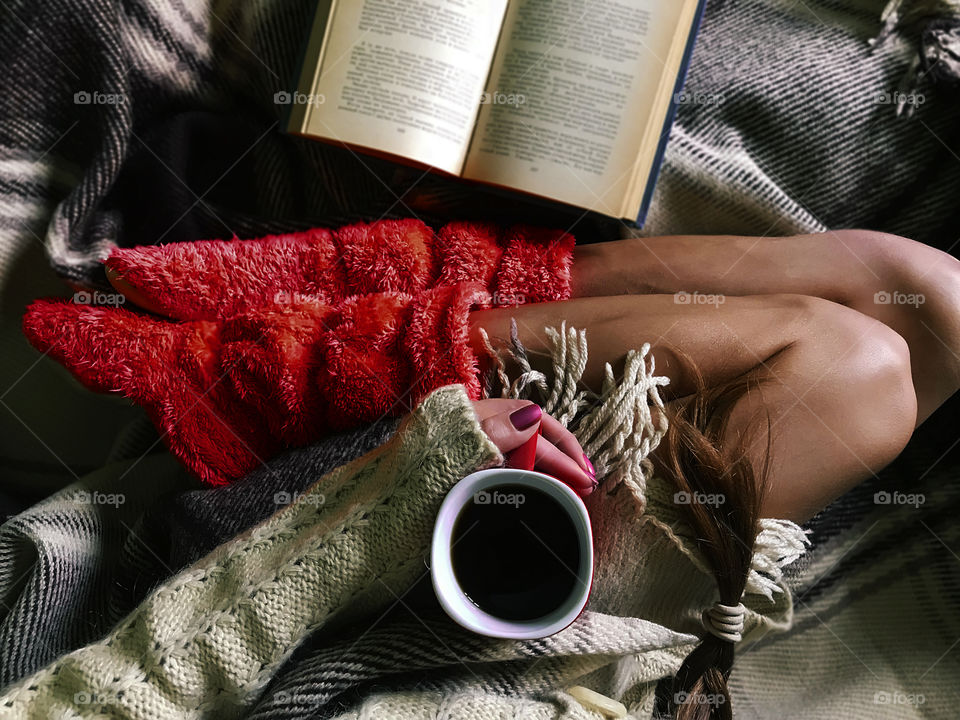 Young woman in red home boots drinking coffee from red cup and reading a book in cozy bed 