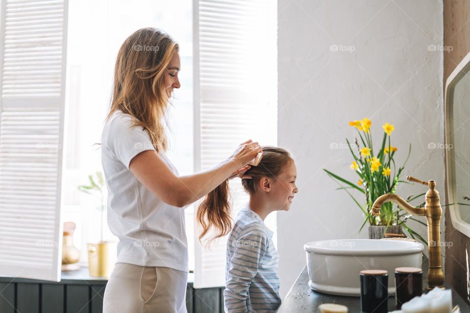 Young mother woman with long hair with little tween girl daughter in pajamas combs her hair on morning in bathroom at home