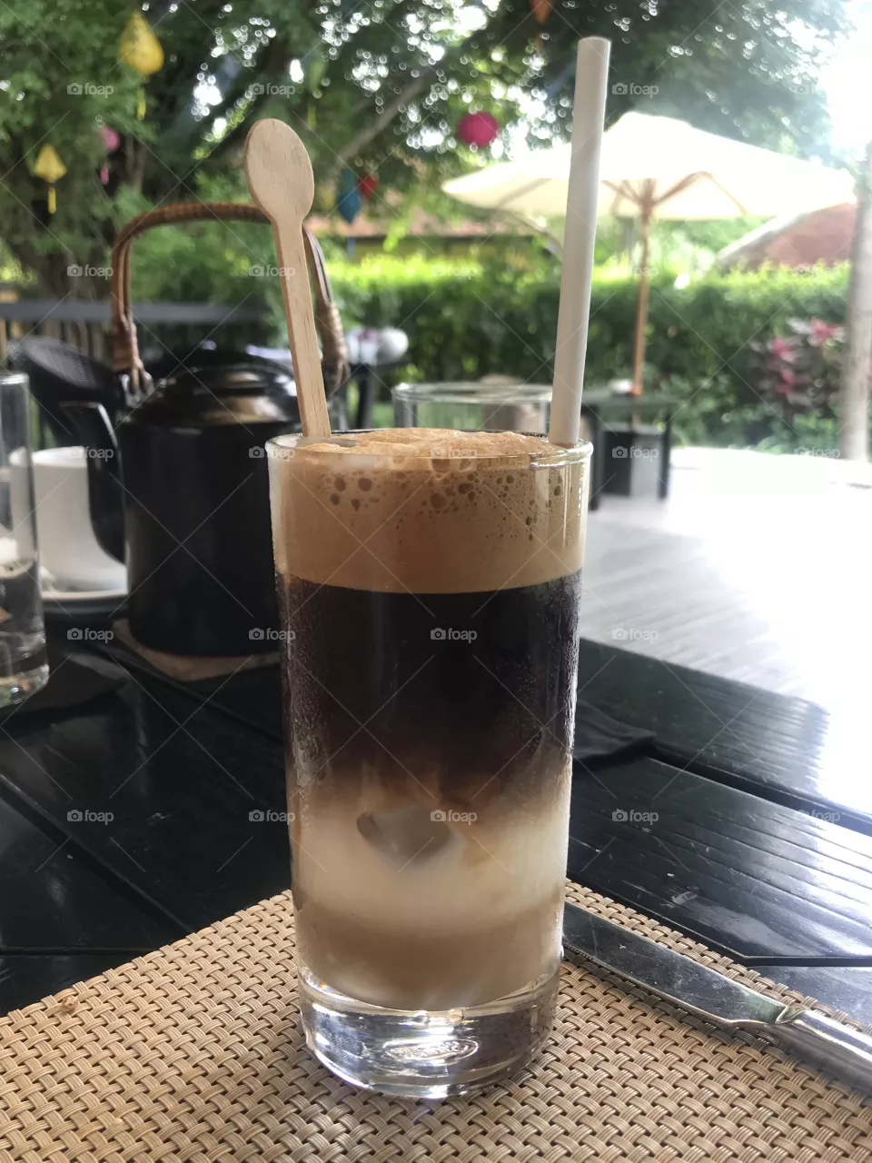 coffee glass on the table,made in 4 color form,black wooden table,1 knife, black tea pot,white cup,glass of water,in the front there are 2 more sets of tables and chairs and a white umbrella.a big tree hanging colorful lanterns, green trees fence