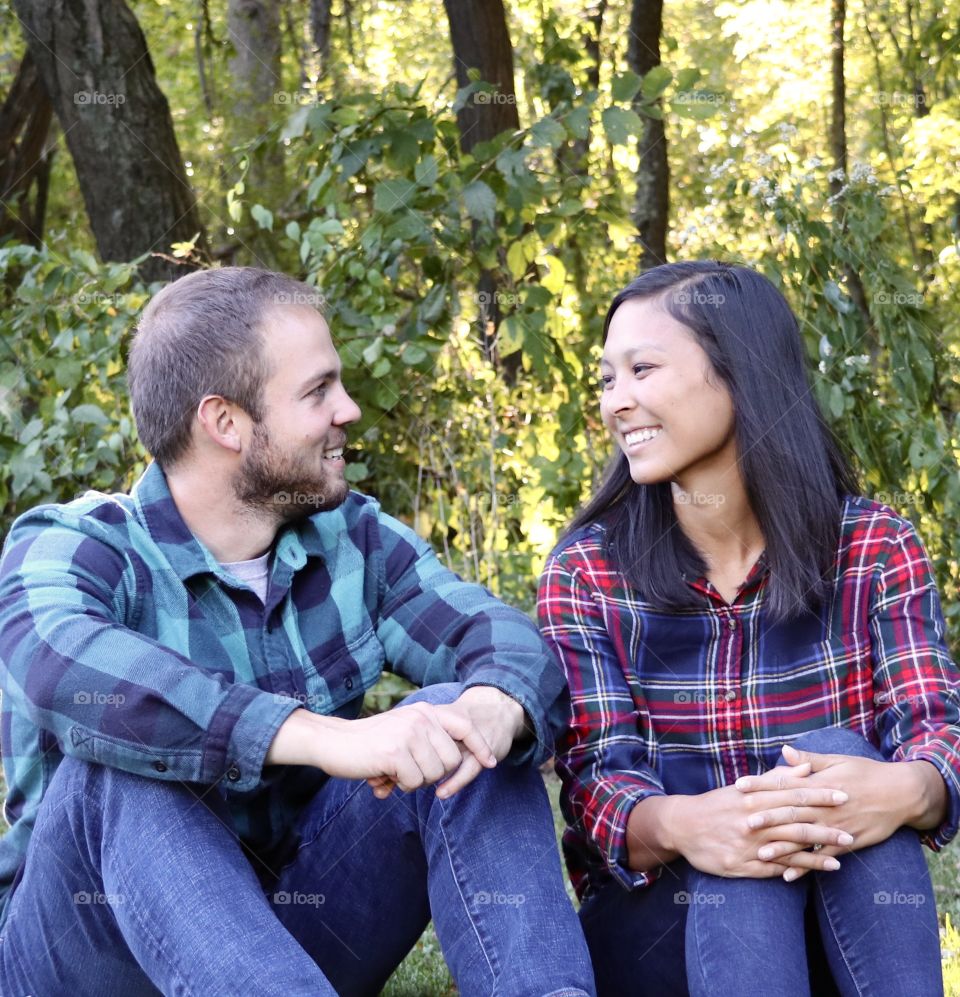 Young man and young lady wearing flannel shirt and blue jeans sitting side by side smiling at each other